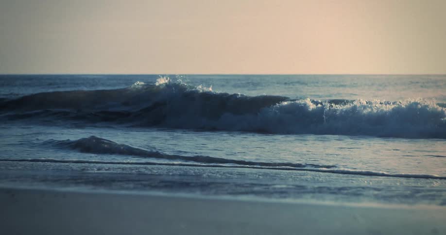 Gentle blue waves roll in one by one breaking with white foam onto the shore under a pale hazy sky