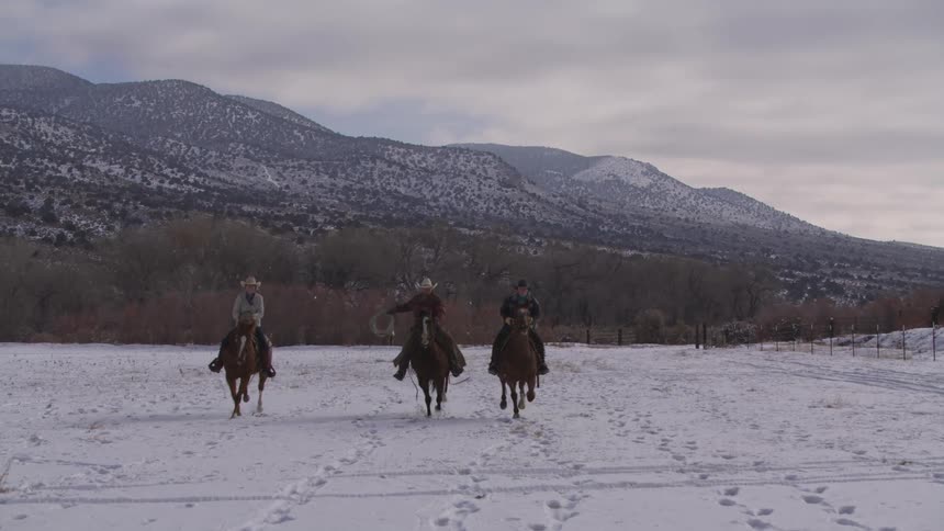 Three riders cross a snowy field surrounded by mountains