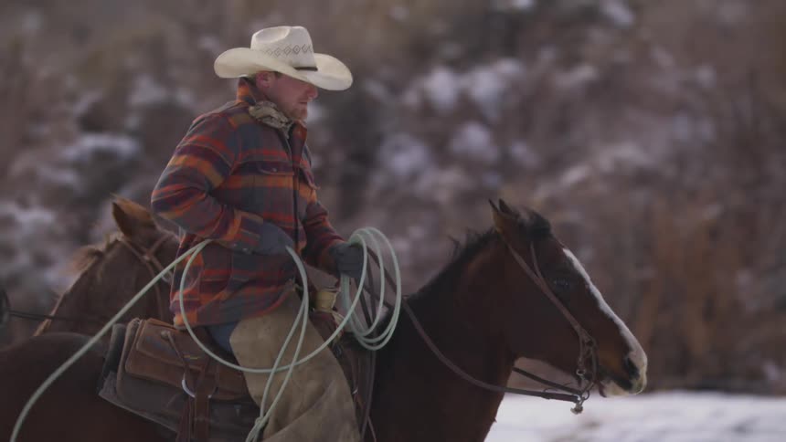 A bearded man dressed as a cowboy in a white hat and plaid jacket rides a brown horse through a snowy rural landscape while holding a coiled rope