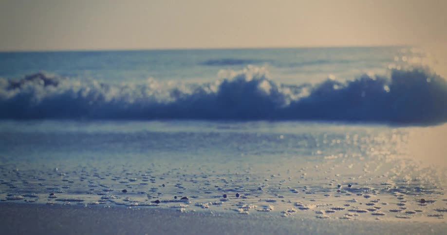 A closeup view shows small waves breaking on a sandy beach under a soft clear sky with the light suggesting early morning or late afternoon the water is a deep blue and white while the sand is a pale slightly golden color giving the scene a serene