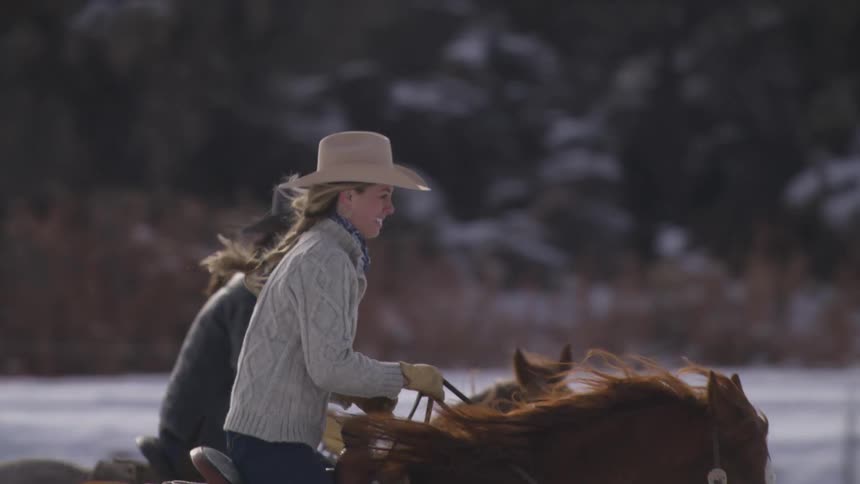 Two women wearing cowboy hats ride their brown horses side by side across a snowy landscape with one woman in a light cable knit sweater and the other in a dark jacket