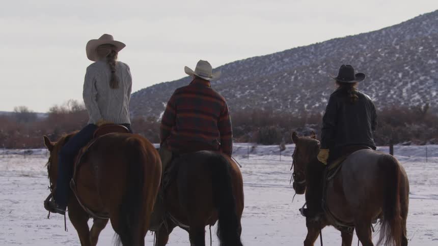 Three people are riding horses across a snowy field near a mountain