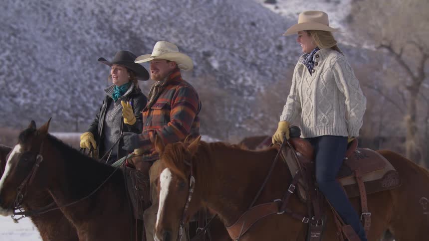 Three people are riding horses through a snowy valley in the mountains