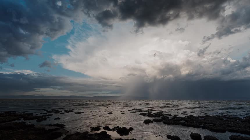 Dramatic coastal scene features dense clouds releasing rain over the sea contrasting with clear sky to the left and rocky foreground