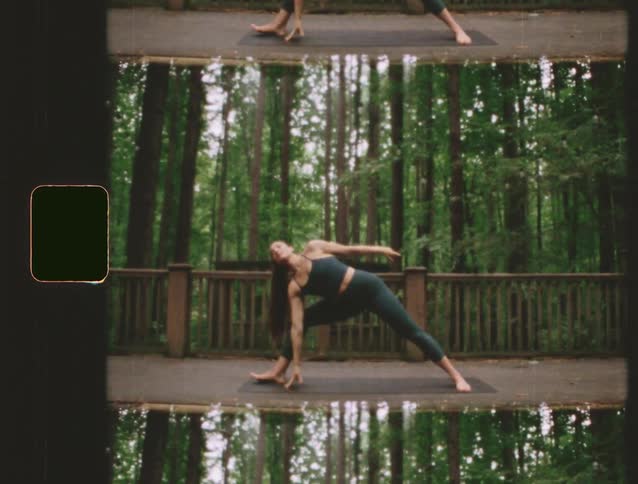 A woman wearing dark workout clothes performs yoga poses on a mat on a wooden deck the background consists of tall trees