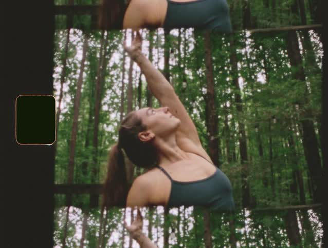 A focused woman wearing dark workout clothing practices a yoga stretch with her arm extended upward on a wooden platform surrounded by tall green trees in a moody forest setting
