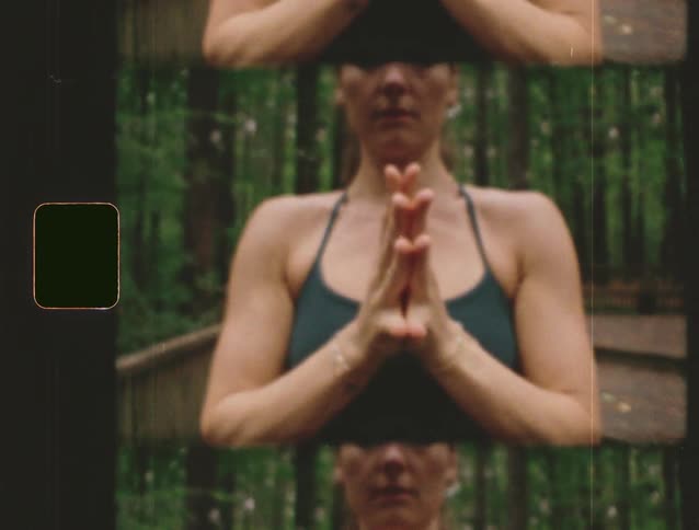 A woman practices yoga outdoors in a forest holding a prayerhands pose in a mirrored kaleidoscopelike visual effect that multiplies her image vertically the footage has an oldfashioned filmlike aesthetic with a visible film sprocket hole on the le
