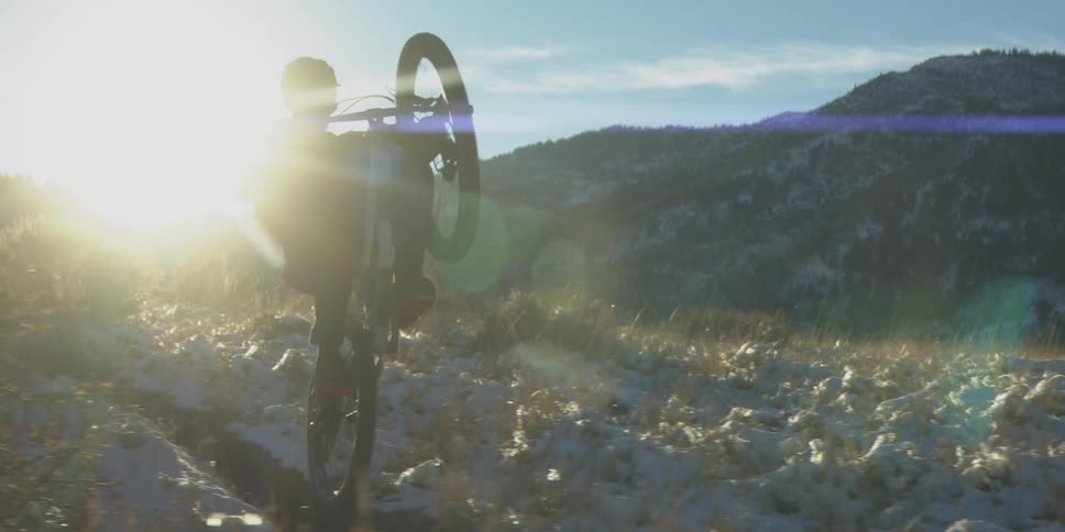 Cyclist performs a stunt on a snowlined trail with low sun among mountains