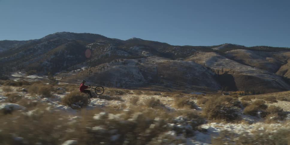 A person riding a bicycle across a field performing a sustained wheelie the rider and bike move horizontally across the frame and large mountains fill the background under a clear sky