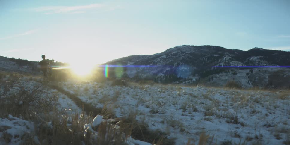 A winter sun bursts over snowdusted grasslands with dark mountains in the distance as a person does tricks on a bike