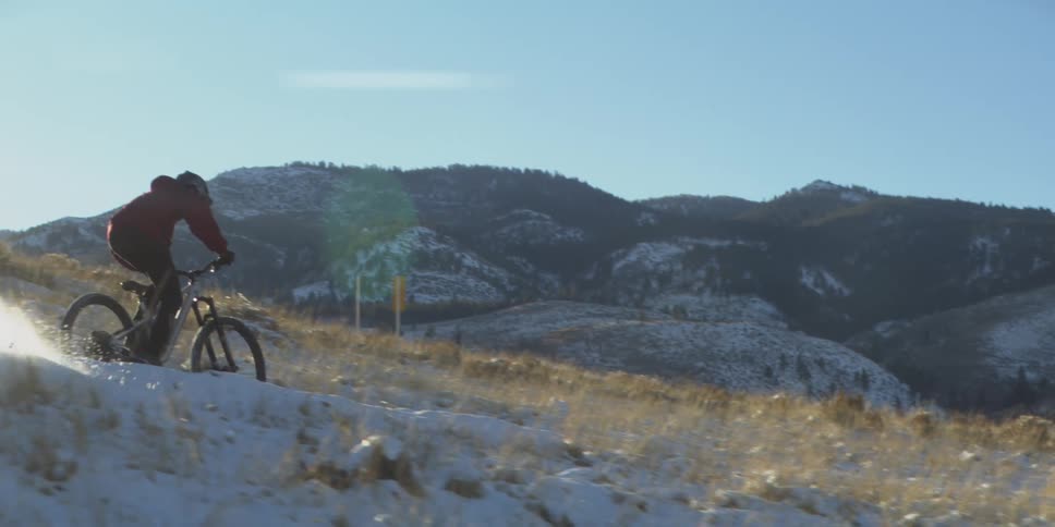 A man in a red jacket and helmet rides a mountain bike up a snow dusted grassy hill then lifts the front wheel into a wheelie against the backdrop of bright sunlight and snow capped mountains