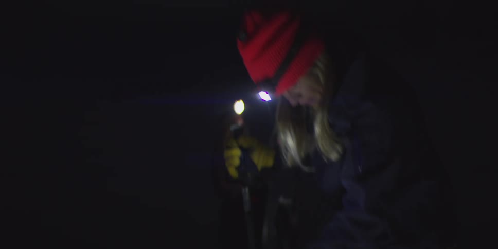 A person wearing ski boots and tan pants adjusts skis in a field of snow at night while other people wearing headlamps stand in the darkness nearby