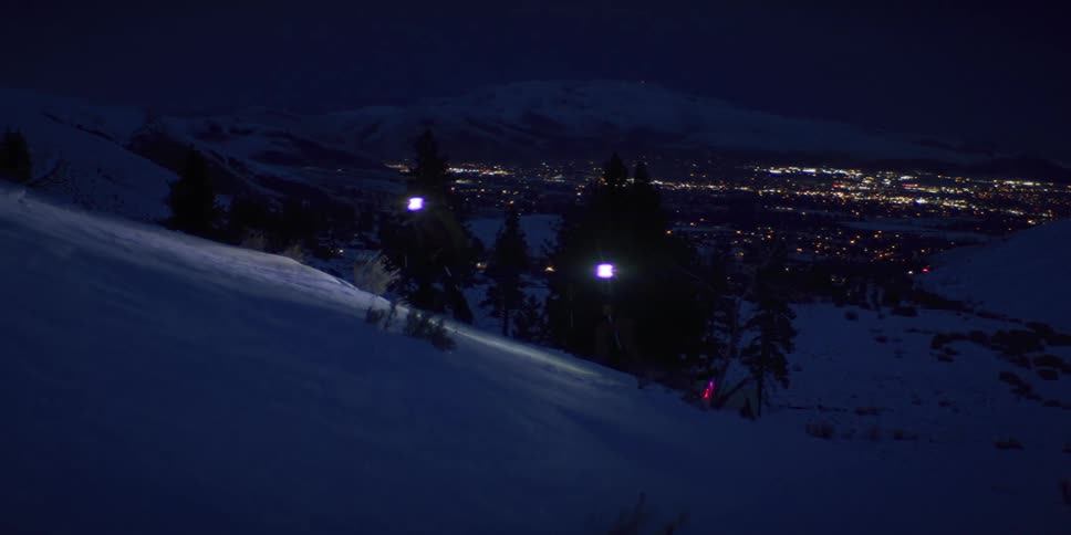 Two skiers wearing headlamps are hiking up a snowy hillside at night with a distant city sparkling below in the valley and a dark mountain visible behind the lights