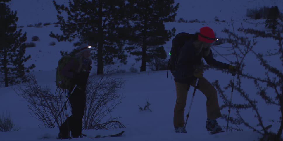 Two backcountry skiers a man and a woman wearing red and white beanies with headlamps and large backpacks stand on a snow covered hill at night under the light of their lamps
