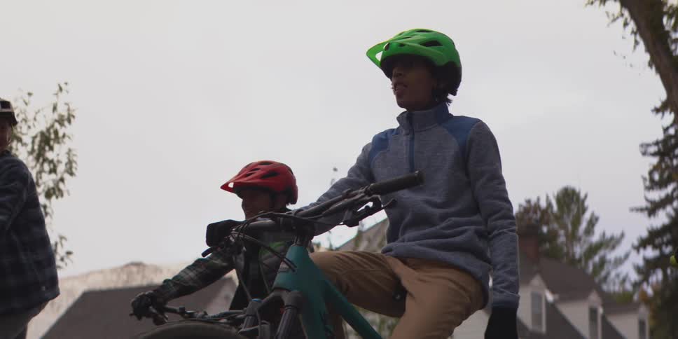 Three children wearing helmets and gloves are riding their mountain bikes on a residential street lined with trees and houses