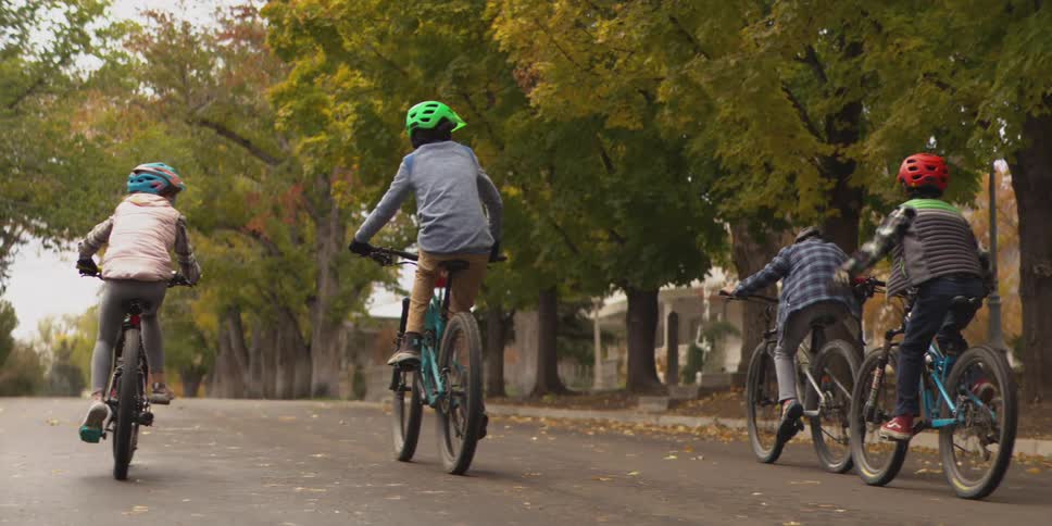 Four children wearing helmets ride bicycles away from the camera on a residential street the street is lined with large trees with green and yellow leaves and a red stop sign is visible on the left side of the frame