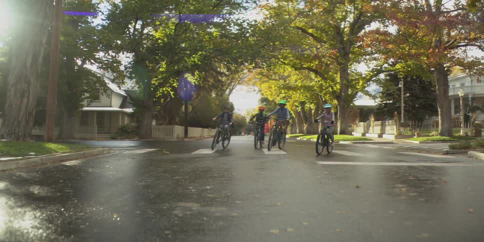 Four young people wearing helmets ride bicycles down a wet residential street that is lined with bright green and autumn trees and large suburban homes