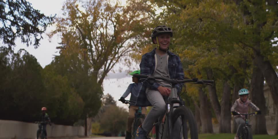 A group of kids ride bicycles down a quiet neighborhood street in autumn with a smiling child in front wearing a helmet