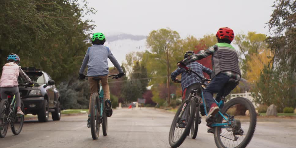 The video is an overtheshoulder shot showing four children riding bicycles down a suburban street all wearing helmets for safetythe setting appears to be late autumn with a mix of green yellow and red foliage and in the distance a snowcapped m