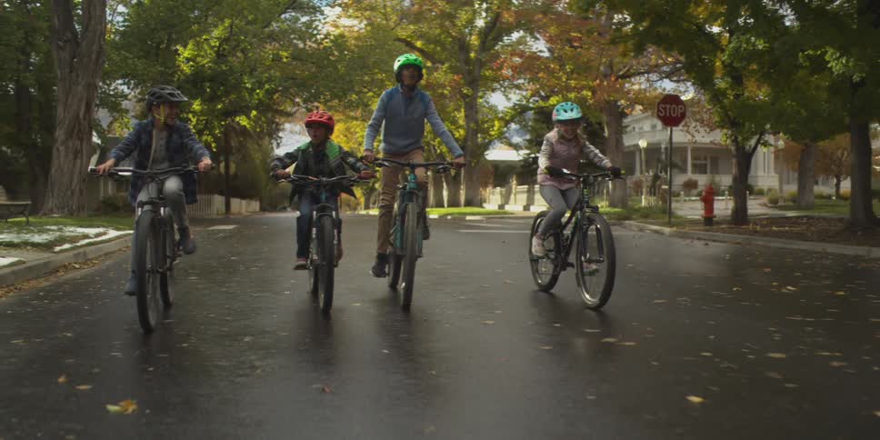 Four kids ride their bikes side by side in the middle of the street in a suburban area at midday