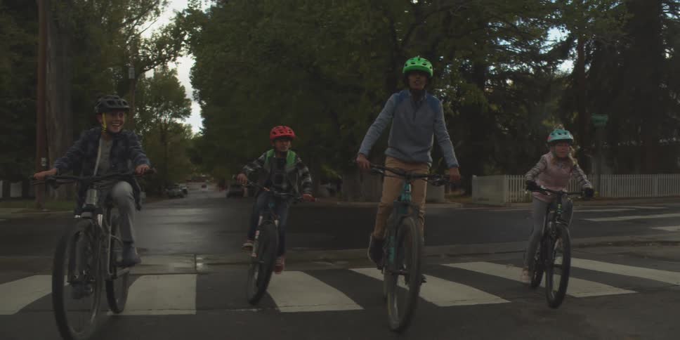 Four kids ride their bikes in the middle of a rural street at midday wearing helmets