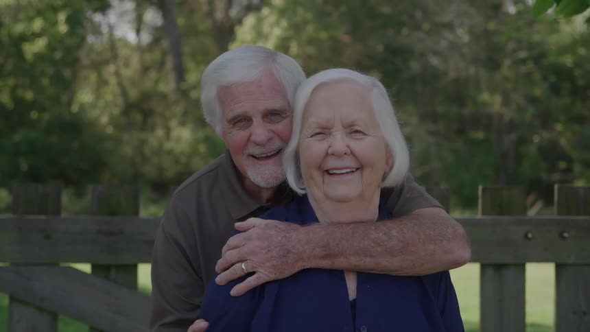 An elderly couple embraces and smiles warmly outdoors in soft natural light