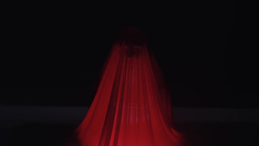 A ghostlike figure stands in darkness beneath a flowing red veil softly lit from below