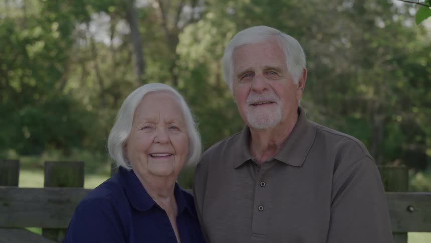 The video is a static medium shot of a happy elderly couple standing sidebyside both smiling directly at the camera they are outdoors with a wooden fence and a background of blurry green trees and foliage