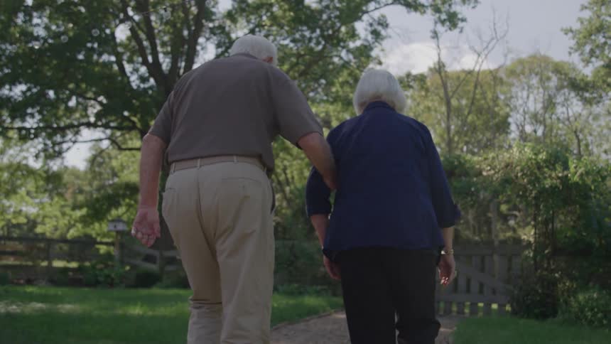 The video is an overtheshoulder shot from behind an elderly couple walking away from the camera along a path the man has his arm around the womans shoulder as they stroll through a sunny green outdoor setting011