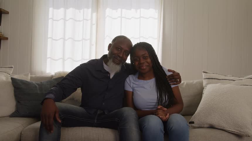 A smiling man has his arm around a young woman as they sit together on a couch both looking forward in a sunlit living room