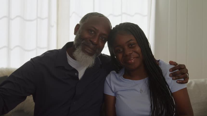 The video is a medium closeup of an older man and a young woman sitting closely together possibly a father and daughter or grandfather and granddaughter they look at each other and then both turn to smile warmly at the camera with the mans arm around