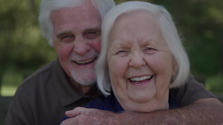 The video is an extreme closeup of a smiling elderly couple embraced looking directly into the camera the man stands behind the woman with his arms wrapped around her and both have wide joyful expressions and show a lot of laughter and happiness