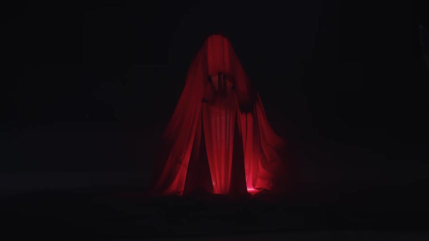 A dark figure hides behind flowing red fabric in dramatic backlight