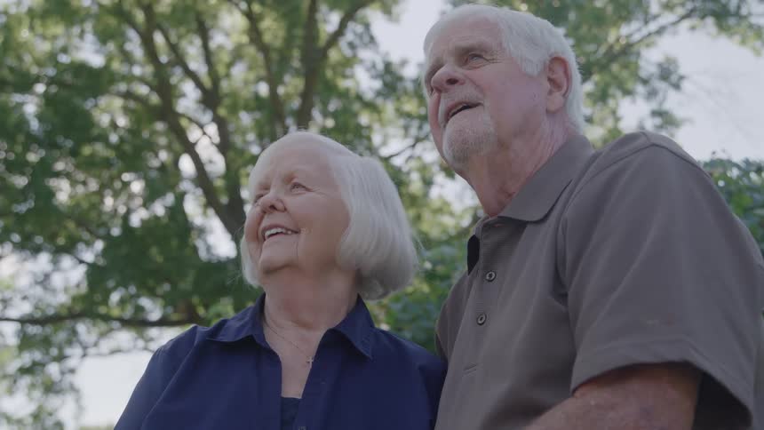 A smiling elderly couple stands close together outdoors under leafy trees on a bright day