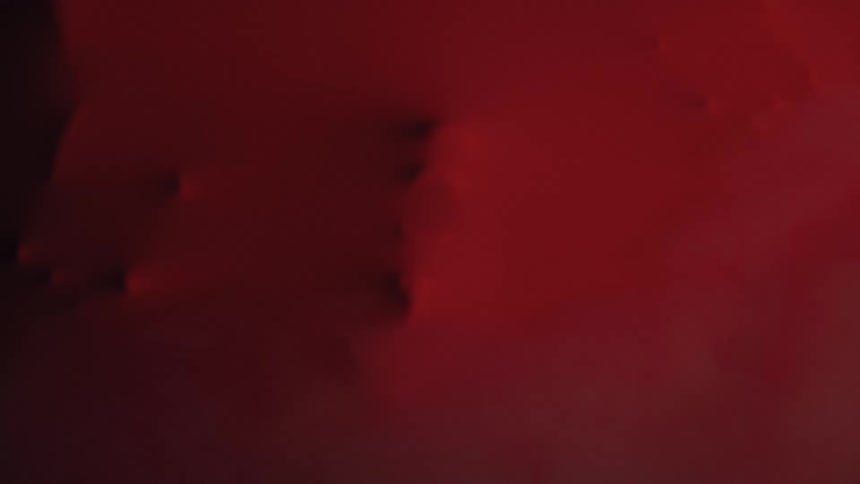 Hands and other forms press and push against a stretchy dark red latex barrier creating distorted struggling shapes from behind the material
