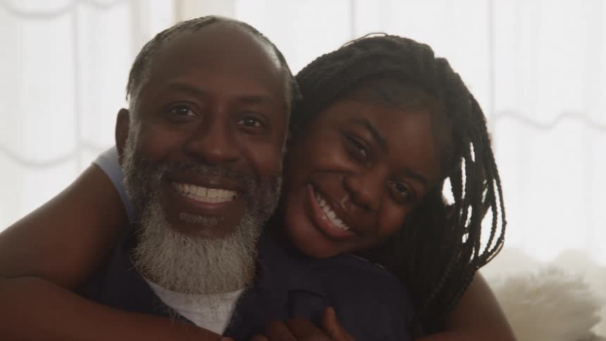 A smiling man with a gray beard is hugged from behind by a cheerful woman with braids both beaming in soft window light