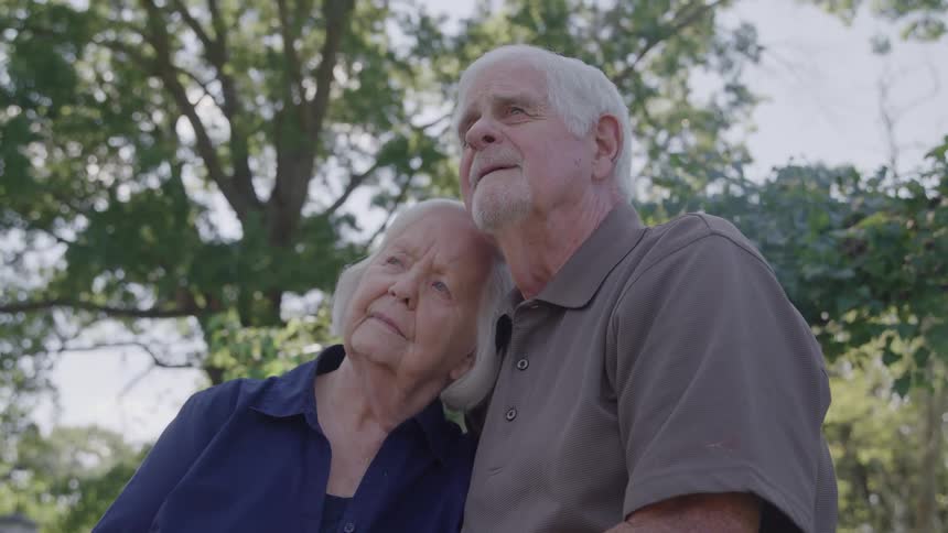 The video is an intimate shot of an elderly couple outdoors with the woman resting her head gently on the mans shoulder they are both looking up and away from the camera standing beneath a large leafy green tree in soft daylight011