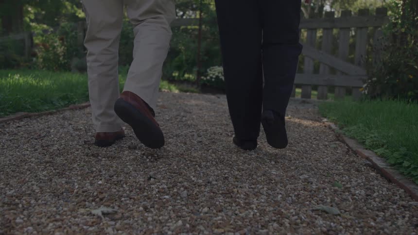 A lowangle shot follows the legs and feet of two people as they walk sidebyside away from the camera down a sunlit gravel garden path