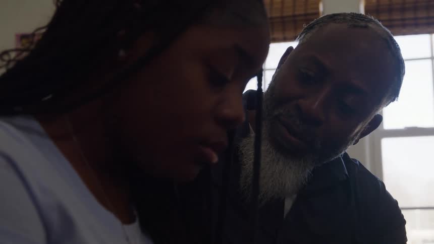 The video is a closeup shot of an african american man and a young woman with braids looking down with concerned and somber expressions the man who has a white beard is seated next to her indicating a moment of serious or difficult conversation in a