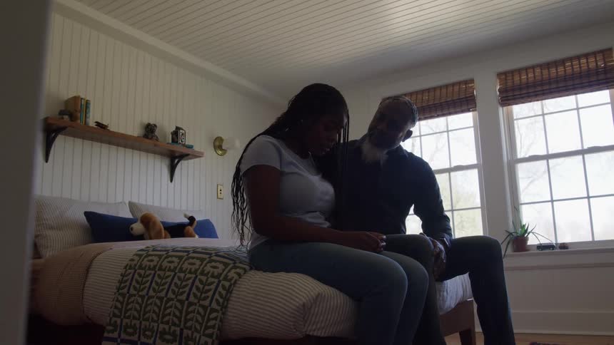Father sits on a bed next to his daughter and comforts her