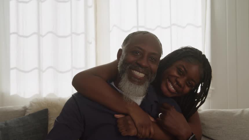 A daughter leans down to hug her father seated on a couch
