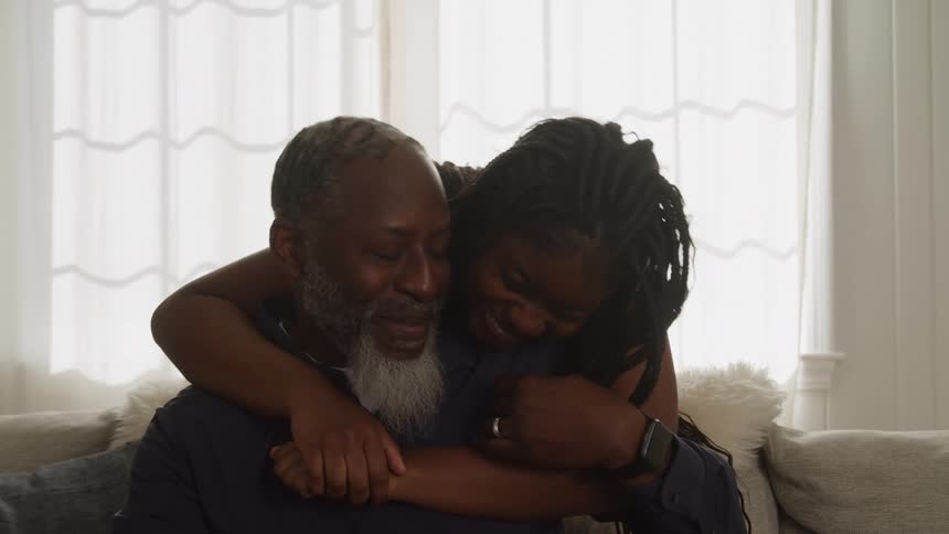 The video shows an elderly black man with a gray beard and a younger black woman embracing and smiling while sitting on a couch indoors the woman is standing behind the man with her arms wrapped around him and they both look genuinely happy and affectio