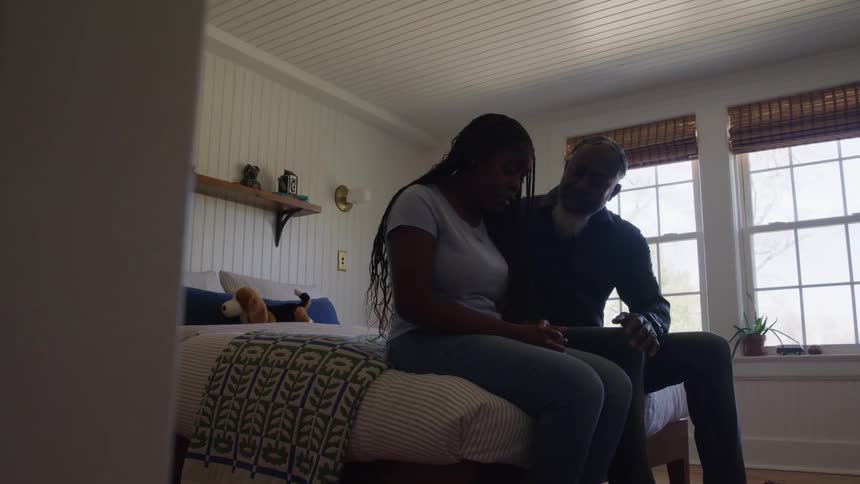 Father and daughter sit together on the edge of the bed in a sunlit room