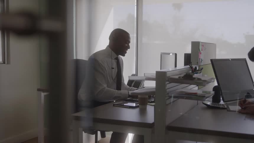 A man talks on the phone and works on computer at his desk in an office
