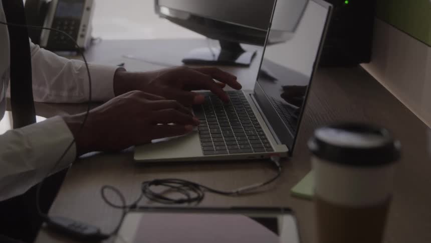 A man in a white shirt and tie types on his laptop at a cluttered office desk with a coffee mug and smartphone nearby