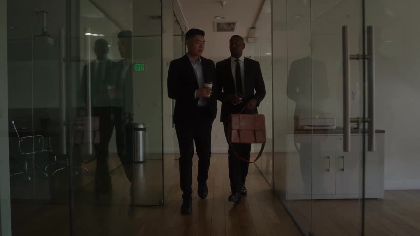 Two professionals walk through modern office hallway with coffee cups and casual conversation