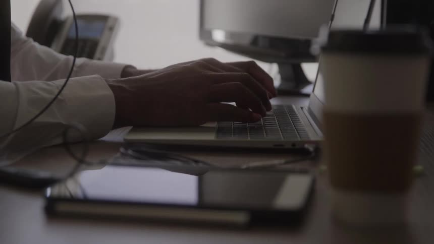 A persons hands type on a laptop at a desk with monitors a tablet and a takeaway coffee cup