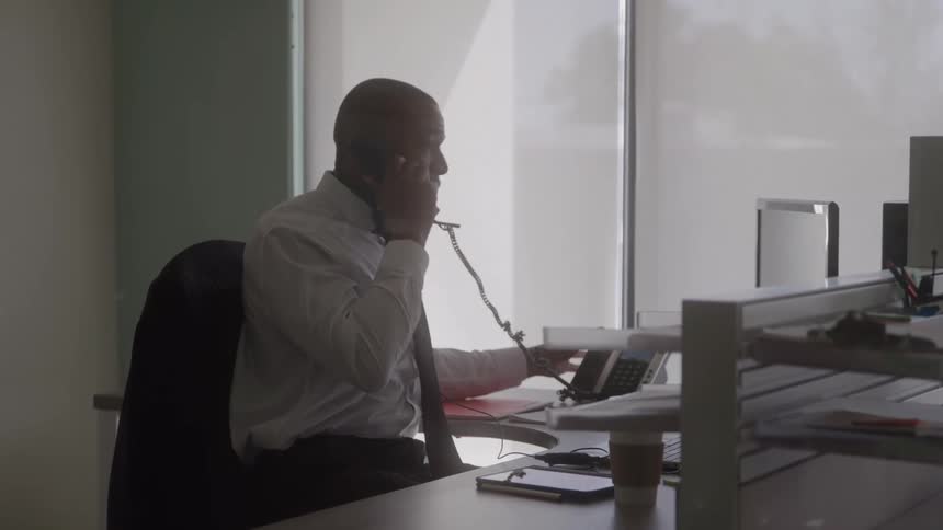 A businessman in formal wear talks on a corded phone at his desk in a bright office