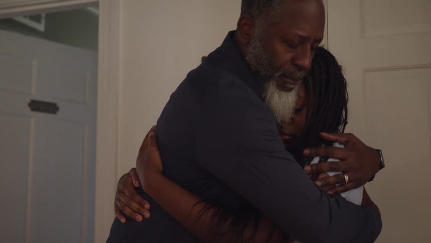 Father and daughter standing indoors with white wall and door in the background embracing