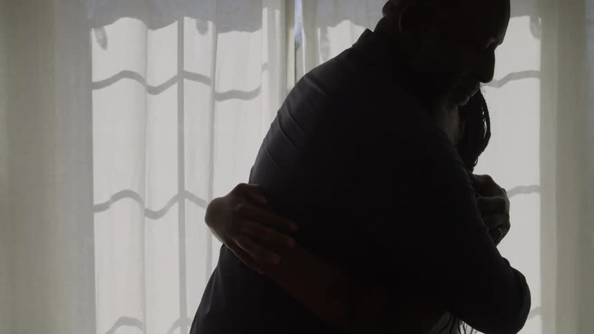The video is an interior shot of a man and a young woman standing in front of a brightly lit window creating a strong silhouette of their figures the man who has a white beard is seen embracing and comforting the young woman who has her arms crossed
